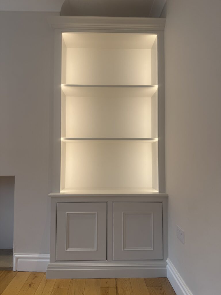 Alcove unit painted pale grey with lit open shelves above panelled cupboards
